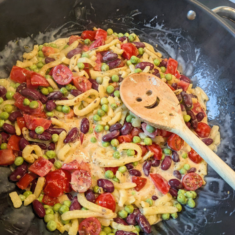 Eine bunte Gemüsepfanne mit Erbsen, Kidneybohnen und veganen Spätzlen. Der Holzkochlöffel hat ein lächelndes Gesicht.